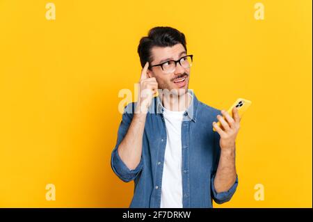 Confused puzzled caucasian unhaved guy, stylishly dressed, holding a smartphone in his hand, looking pensively to the side, standing an isolated orange background Stock Photo