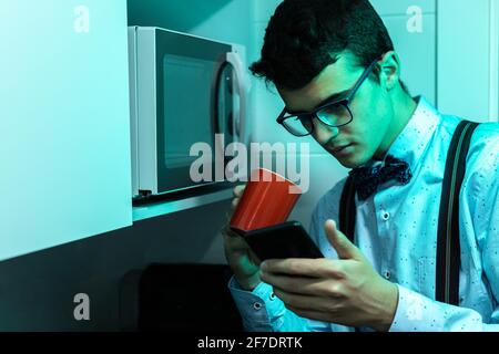 Young man in a kitchen drinking a red cup of coffee with milk and looking at the messages on the mobile phone while having breakfast and wearing a sui Stock Photo