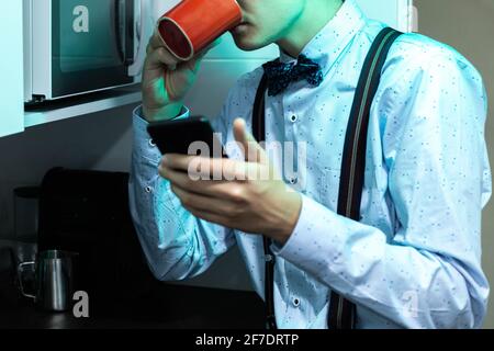 Young man having a red cup of coffee with milk for breakfast, wearing a suit and bow tie and using the mobile phone to view and send messages on the i Stock Photo