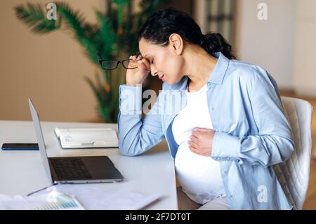 Upset tired pregnant mixed race woman, business lady, manager or freelancer, uses laptop for work, overworked with business projects, under stress, taking break, taking off glasses, closing eyes Stock Photo