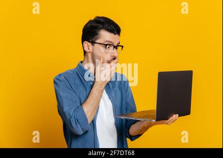 Confused puzzled stylish unshaven caucasian guy with glasses holding an open laptop in his hand, looks surprised at it, covering mouth with hand, while standing on isolated orange background Stock Photo