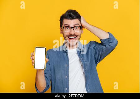 Amazed shocked surprised caucasian guy in denim shirt and glasses holds smartphone with blank white screen for your text, touches head with hand, stands on isolated orange background, looks at camera Stock Photo