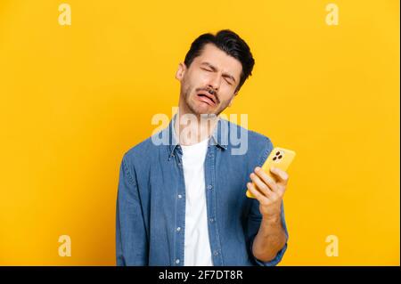 Caucasian stylish guy holding a smartphone in his hand, with a negative unhappy facial expression, received a negative message, or an unwelcome call, stands on an isolated orange background Stock Photo