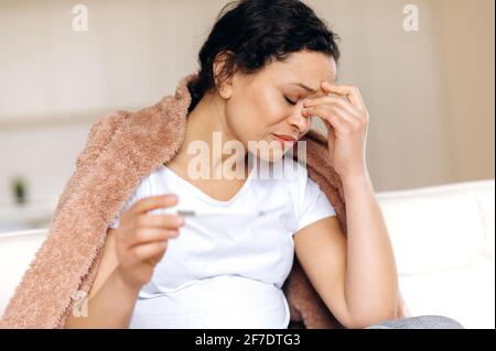 Exhausted sick pregnant mixed race woman sitting upset at living room on couch under blanket, massage her eyes with hand while taking temperature with thermometer, she has a cold, flu Stock Photo