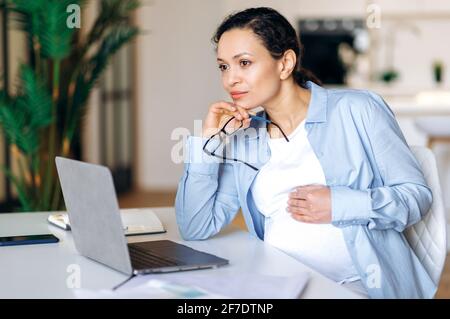 Focused smart confident beautiful pregnant mixed race woman working on laptop at at workplace, taking break, thinking about business project or vacation, looking away, takes off glasses,stroking belly Stock Photo