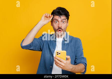 Amazed handsome unshaven caucasian guy holding smartphone in his hand, looking at the camera in surprise with his glasses raised, shocked facial expression, isolated orange background Stock Photo