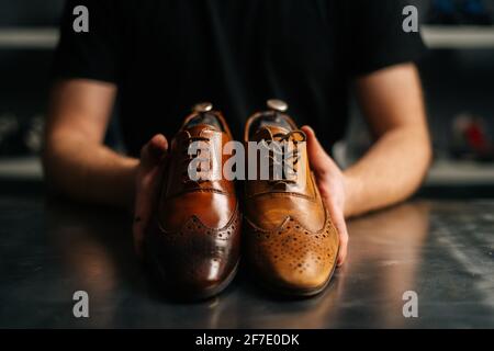 Close-up hands of male shoemaker holds old light brown leather shoe and repaired shiny shoes after restoration working. Stock Photo