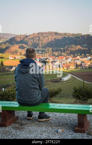 Back view of a man sitting on a bench and resting while looking towards the village he used to know. Stock Photo