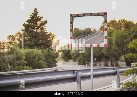 Rectangular road mirror with red and white frame on a curve on a road. Hazardous turn where a mirror is in a good aid. Sunny evening with no cars.