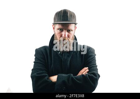 Angry looking hipster man with a beard and a baseball hat wearing a black winter coat on a white studio background. Angry bearded man. Stock Photo