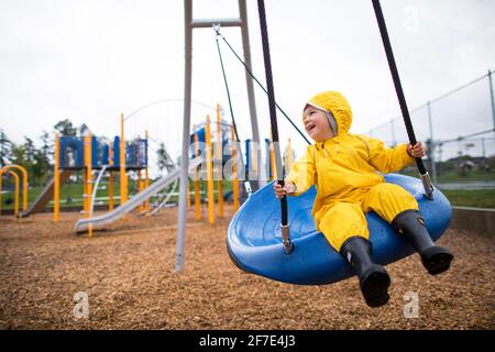 Happy toddler boy rides on swing at the park on a wet day Stock Photo