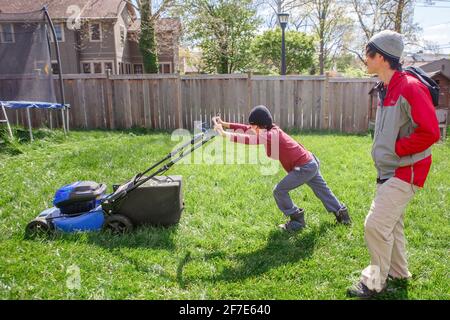 Boy works hard to push electric lawn mower while father watches on Stock Photo