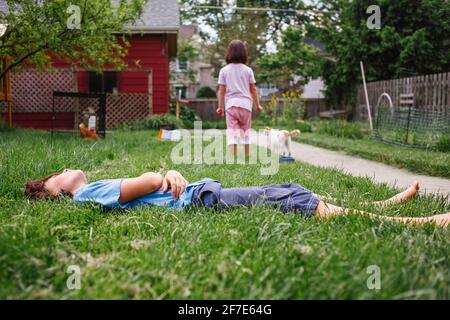 a boy lays in green grass in yard with sister and cat in background Stock Photo
