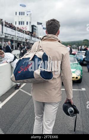 Young stylish race driver heading home after the race .Race driver on the track with a training bag on his shoulder and a building in the background. Stock Photo