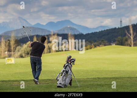 a man hitting a golf ball on a driving range of a golf field. Visible grass flying around and bag with clubs on a beautiful background with mountains Stock Photo