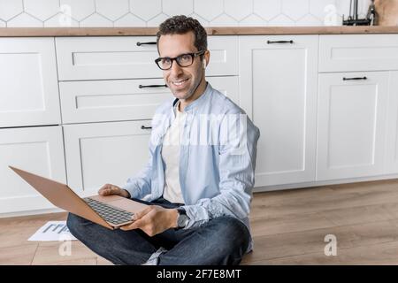 Attractive male freelancer is using laptop, looking at the camera, smiling. Successful stylish businessman working remotely on project or financial Stock Photo