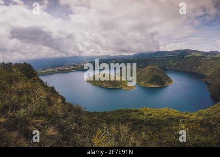 Looking towards the island on famous Lake Cuicocha in Ecuador. Panorama over a blue lake, surrounded with lush greens and white clouds. Stock Photo