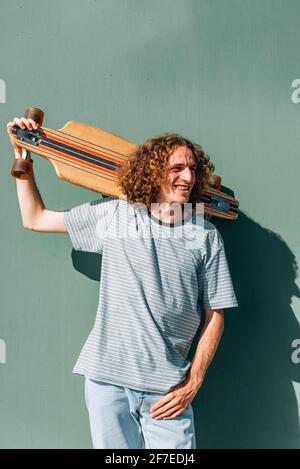 vertical portrait of a young man holding his long board and looking to one side. He has long hair and smiles Stock Photo