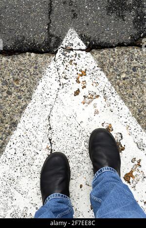 A man wearing jeans and black boots stands on a the tip of a white painted arrow on cracked pavement and concrete Stock Photo