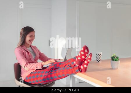 Work online remote from home funny concept. Asian woman relaxing in pajama pants and cozy socks while wearing professional top and suit for videocall