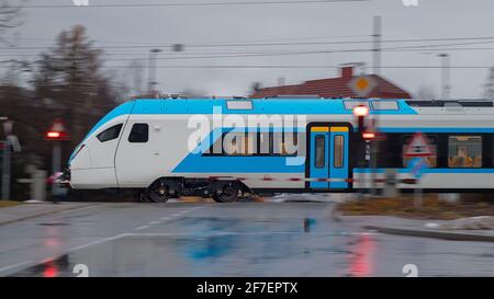 Side view of modern electric passenger train during cold winter rain is rushing through a grade crossing with road with barriers down and red signals Stock Photo