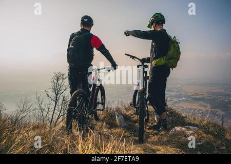 Two men with mountaibikes on a top of the hill. Panorama of a valley with cyclist in the foregroud. One is pointing his fingeer. Stock Photo