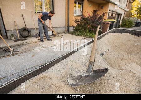 A man is using rakes to straighten and level the sand in front of a residential house. Focus on the shovel in the sand in the foreground. Home constru Stock Photo