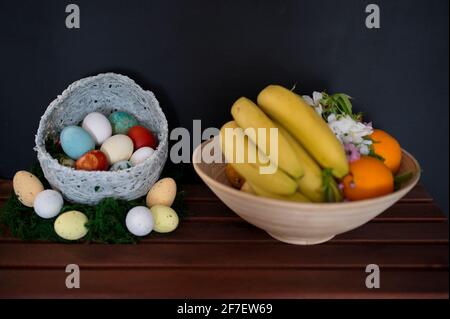 Colorful Easter eggs in wicker basket and bowl with fruits on black background Stock Photo