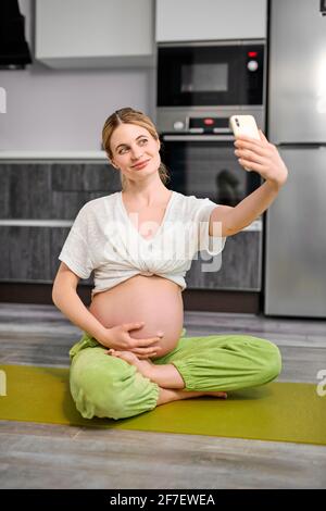 Cute Caucasian Female Take Photo On Mobile Phone, After Doing Yoga Exercises During Pregnancy. Sit On Fitness Mat Smiling, Enjoying Healthy Lifestyle Stock Photo