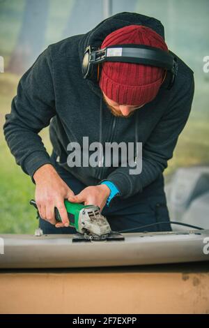 Young service man is using an angle grinder with a wire brush to clean the underside of an old vintage car about to be restored Stock Photo