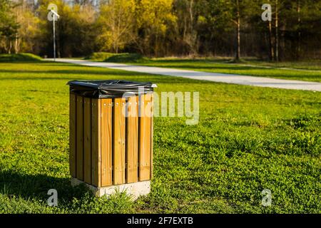 A modern wooden trash can or bin in a park with a black plastic bag visible inside on a sunny day. Walkway or path for pedestrians in the background. Stock Photo