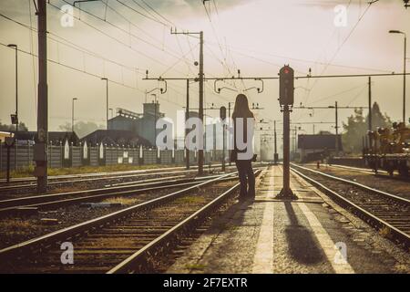 Young woman in coat is waiting for a train and holding a telephone on a narrow station platform between the tracks in early morning hours just after s Stock Photo