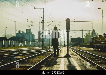 Young woman in coat is waiting for a train on a narrow station platform between the tracks in early morning hours just after sunrise, with fog and mis Stock Photo