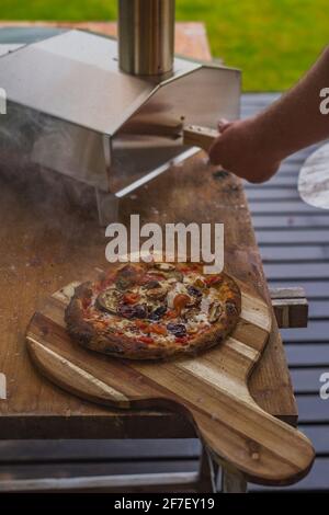Delicious fresh home made pizza on a round wooden plate, directly out of a stainless steel home portble oven fueled by pellets. Outdoor pizza party. Stock Photo