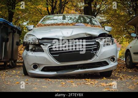 Frontal part of a small city vehicle smashed. Demolished and crashed front of a silver car, parked on a public parking space between the trees and tra Stock Photo