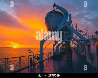 Lifeboat on a cruise ship or ferry at a romantic evening sunset setting. Rescue lifeboats high up on a crane, ready to be deployed. Stock Photo