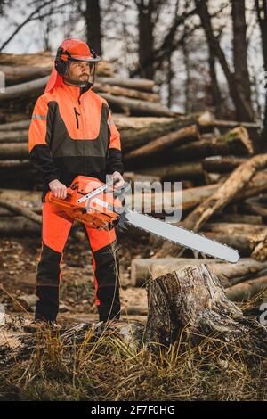 A chainsaw operator standing next to a tree trunk, holding a big orange chainsaw. Protective equipment is used, such as helmet, pants and a vest. Stock Photo