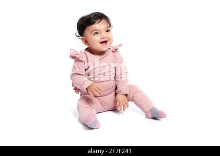 Adorable female baby in lovely pink outfit smiling with mouth open. Attractive little kid sitting on floor and posing, isolated on white studio background. Concept of childhood. Stock Photo