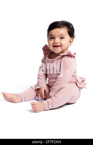 Side view of lovely baby girl in cute pink outfit smiling with mouth open. Attractive little barefoot kid sitting on floor and posing, isolated on white studio background. Concept of childhood. Stock Photo