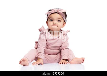 Adorable baby girl in lovely pink outfit and headbandlooking up. Attractive little female kid sitting on floor and posing, isolated on white studio background. Concept of childhood. Stock Photo