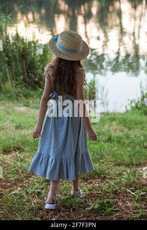 A girl in blue dress and straw hat stands on the shore of the lake Stock Photo