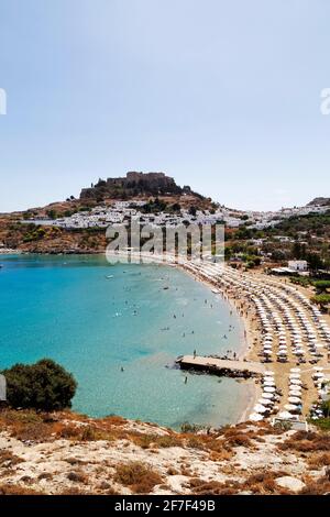 Sunshades are ranged on the beach at Lindos on Rhodes, Greece. The Lindian Acropolis rises over white houses in the fishing village. Stock Photo