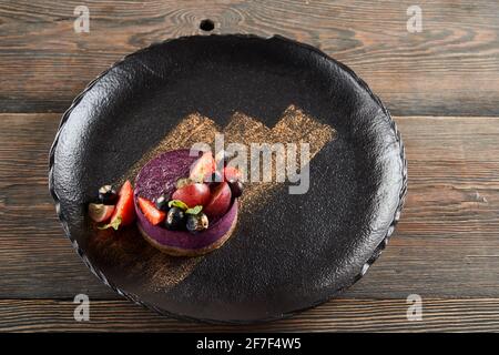 From above view of round piece of traditional blueberry cheesecake served with blueberries and strawberries on wooden table in restaurant. Golden brushstroke decoration on black plate. Stock Photo
