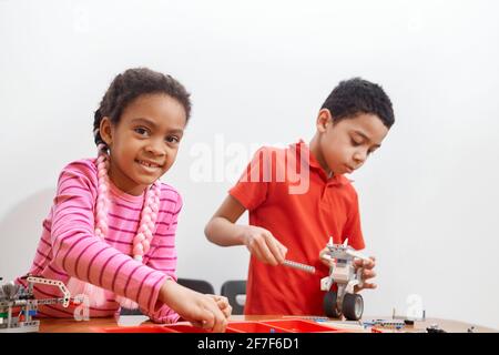 Front view of building kit for group of kids creating toys, having positive emotions and joy, selective focus of girl smiling and looking at camera. Close up of african friends working on project. Stock Photo