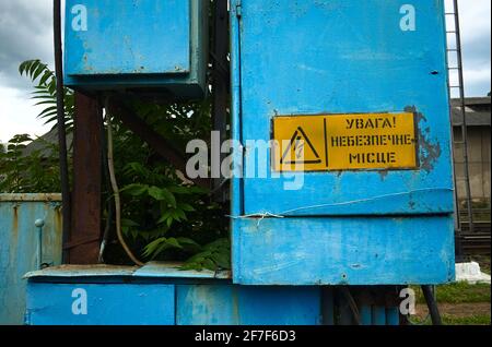 Old high voltage transformer sign on blue metal board. Electric power box near railroad tracks with danger triangle sign and caution notice on yellow Stock Photo