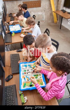 Interesting building kit for kids on table with computers. From above view of boys and girls creating toys. Science engineering. Nice interested friends chatting and working on project together. Stock Photo