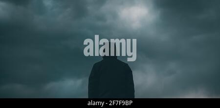 Rear view of male person wearing hooded jacket against dark moody dramatic clouds at sky, man looking into uncertain ominous future Stock Photo