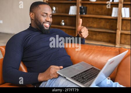 Smiling African-American man talking online via video call on the laptop in a relaxed atmosphere, a black guy lying on the couch and showing thumbs up at webcam in sign of good connection Stock Photo