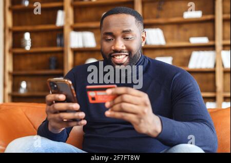 Smiling African-American man shopping online sitting on the sofa at home, holding credit card and smartphone, dark skinned multiracial guy making food order, purchasing online, booking and paying Stock Photo