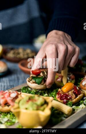 a young caucaisan man takes an appetizer from a wooden tray with some different vegan appetizers, with different toppings, placed on a gray rustic tab Stock Photo